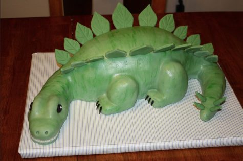 A dinosaur cake sits on a stand, ready to be eaten.